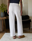 Rear view of a person in Frank & Eileen's Bella Sweatpants with brown shoes, standing on a light rug. A dark sofa and side table with a vase of greenery complete the cozy background setting.