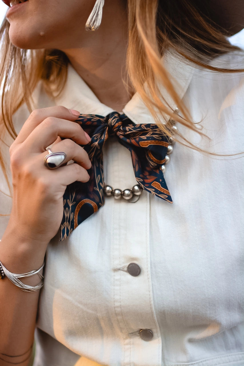 A woman wears a cream-colored, wrinkle-resistant button-up shirt with Faire’s Western Harvest Twilly scarf as a bow and a silver beaded necklace. She accessorizes with a large ring, bracelet, tassel earrings, and her light brown hair peeks through.