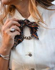 A woman wears a cream-colored, wrinkle-resistant button-up shirt with Faire’s Western Harvest Twilly scarf as a bow and a silver beaded necklace. She accessorizes with a large ring, bracelet, tassel earrings, and her light brown hair peeks through.