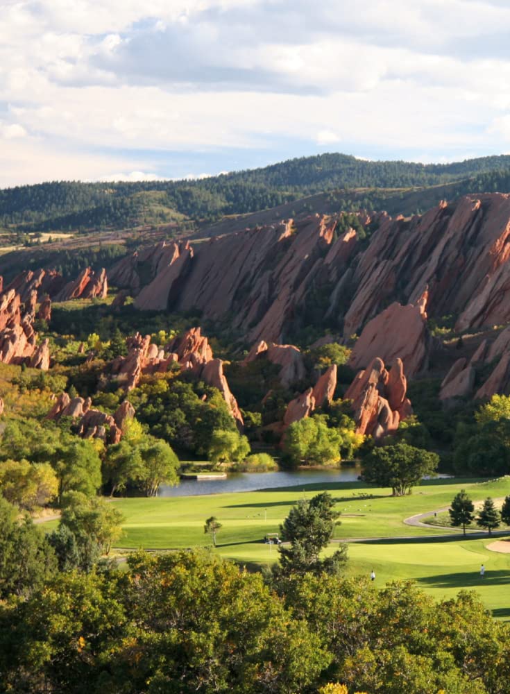 Experience dramatic red rock formations, lush trees, and a top golf course beside a pond—featured in "150 Golf Courses You Need to Play" by Simon & Schuster Inc.—all set against rolling hills and a partly cloudy sky.