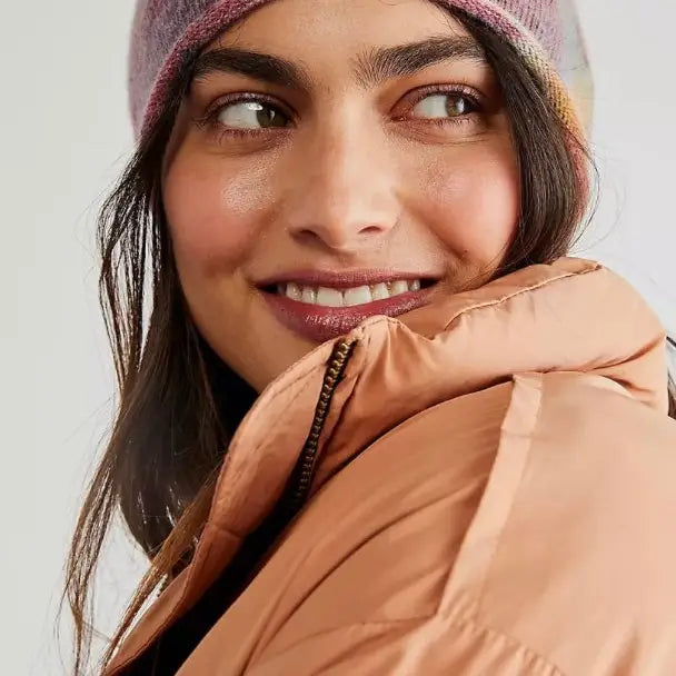 A woman in a colorful Faire Cashmere Beanie and a peach-colored puffer jacket gazes to the side with a gentle smile.