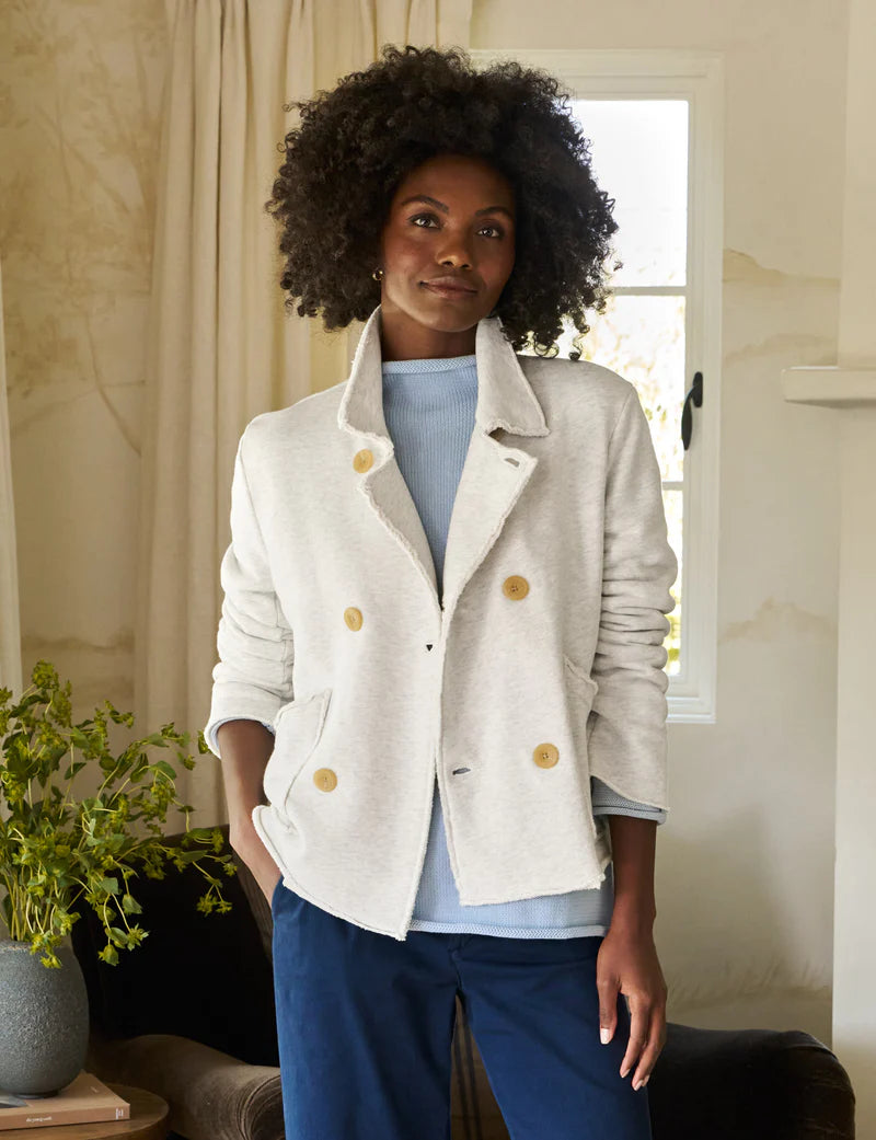 A person with curly hair stands near a window with light curtains, wearing a Frank & Eileen MINI BELFAST Crop Peacoat over a blue top and dark pants. A plant is visible in the background.