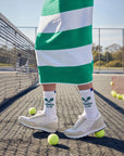 A person wearing vintage-inspired white sneakers, Kule’s The Women's Tennis Sock WHT, and a green-and-white striped skirt stands on a tennis court with scattered tennis balls.