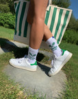 A person in white sneakers and Kule's The Women's Tennis Sock WHT stands on a stone path, holding a green-and-white striped tote bag, with grass and trees in the background under bright sunlight.