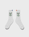 The Women's Tennis Sock WHT by Kule features vintage-inspired white socks with "KULE Tennis Club" and two green tennis rackets. One sock has a blue tab, the other an orange tab at the top for a playful touch.