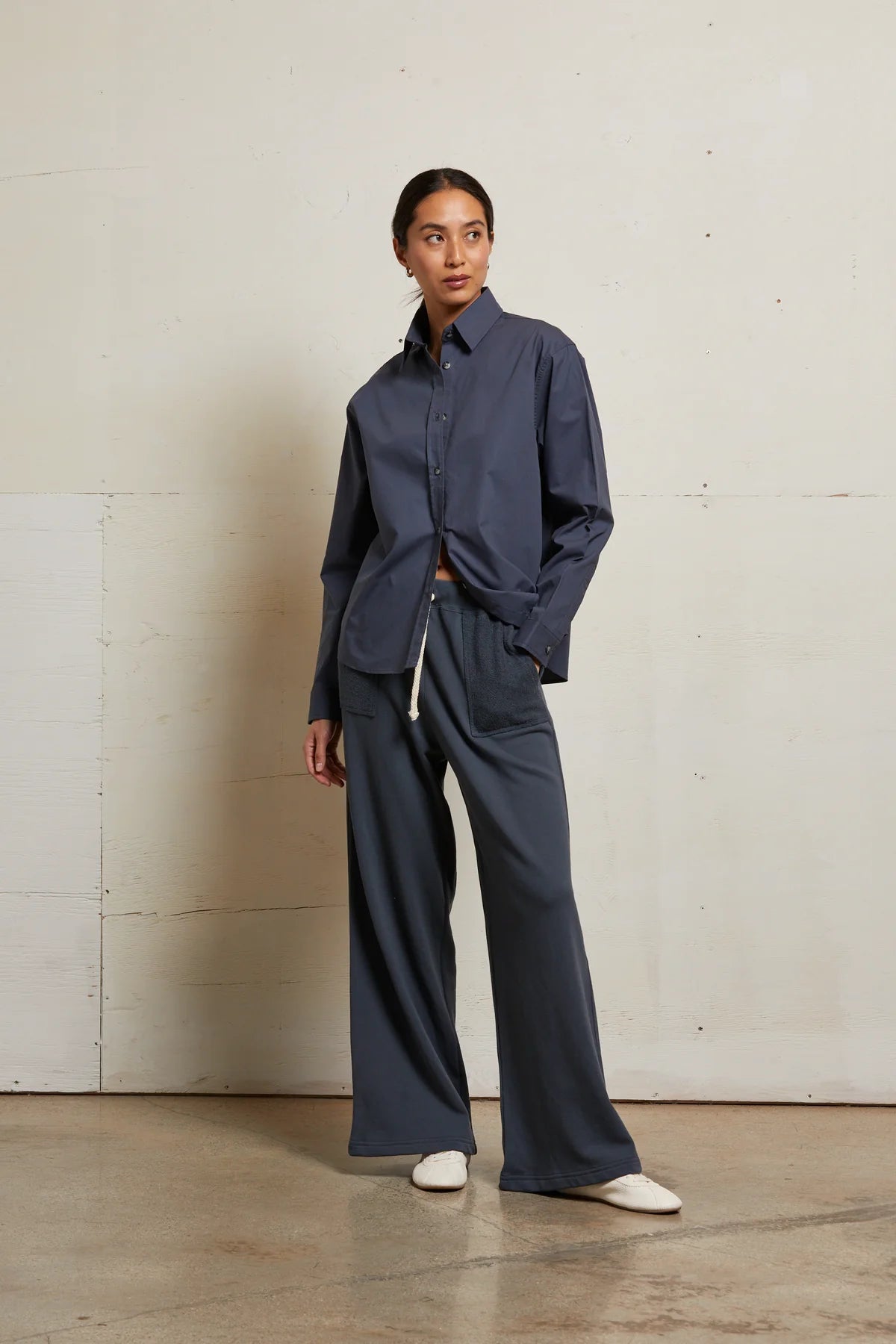A woman stands indoors against an off-white wall, wearing Perfectwhitetee’s French Terry Pant and a loose dark blue button-up shirt with white shoes. She looks to the side with one hand in her pocket.