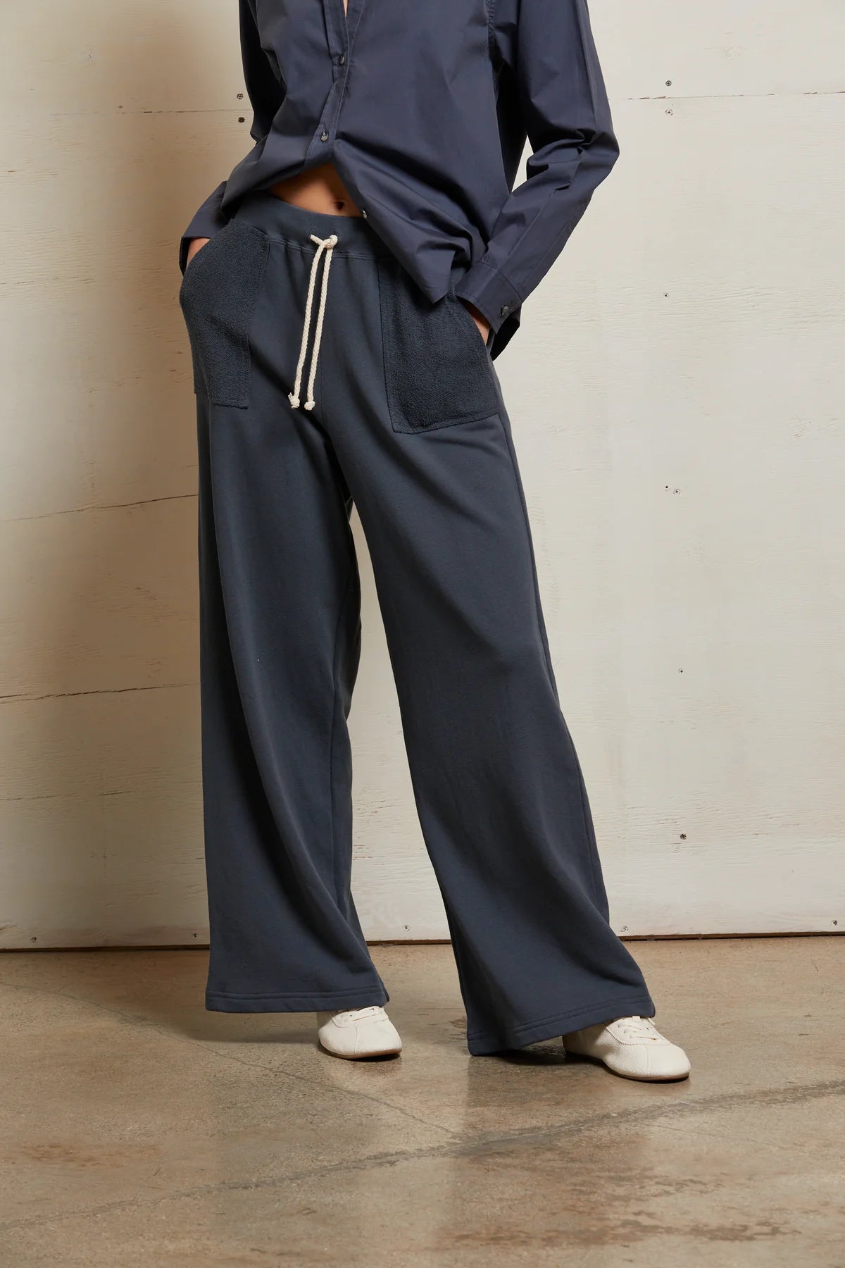 A person wears the Perfectwhitetee French Terry Pant in navy—loose, straight-leg, 100% cotton with patch pockets and a drawstring waist—paired with a navy shirt and white sneakers on a concrete floor.