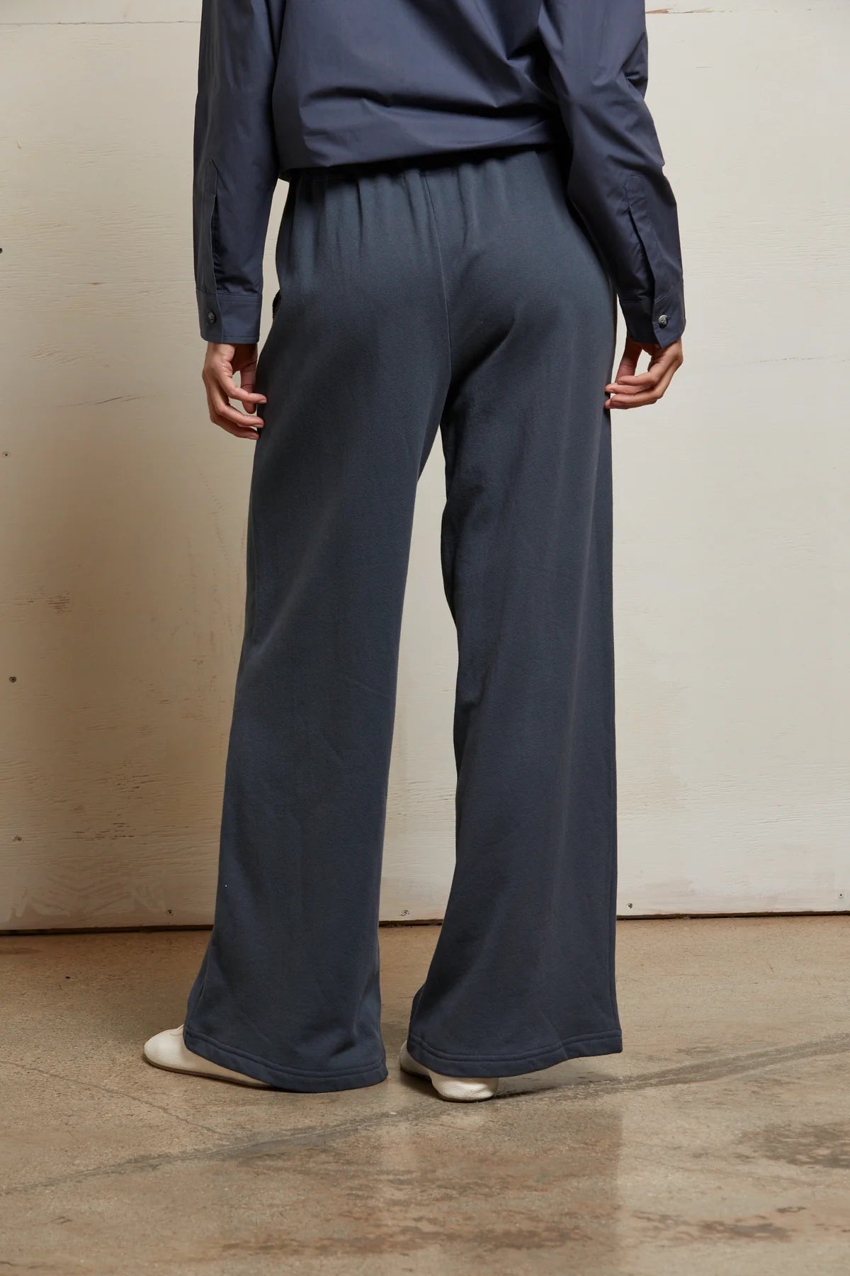 A person stands indoors on a concrete floor, back to the camera, arms relaxed, wearing Perfectwhitetee's loose-fit dark gray French Terry Pant and matching long-sleeve shirt, both crafted from 100% cotton French terry.