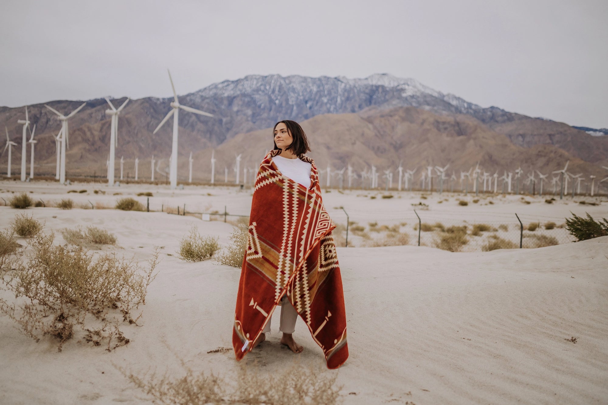 A woman stands on sandy desert ground wrapped in the Faire Achuar Blanket, with wind turbines and mountains in the background under an overcast sky.