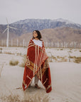 A woman stands on sandy desert ground wrapped in the Faire Achuar Blanket, with wind turbines and mountains in the background under an overcast sky.