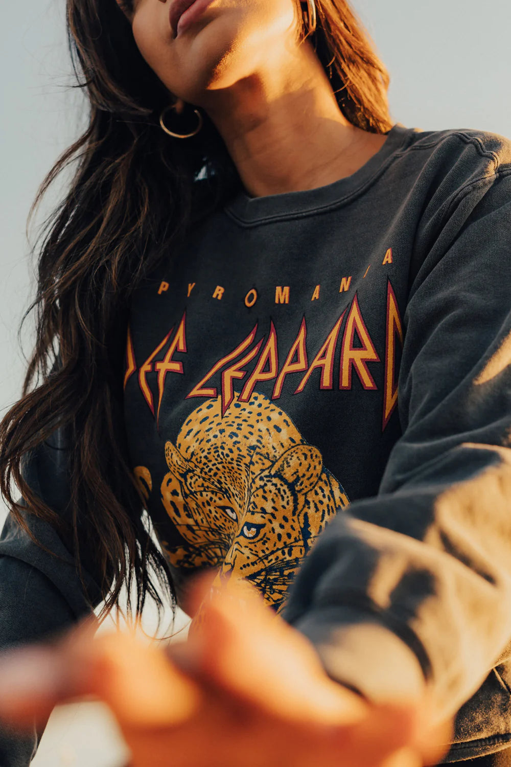 A person dressed in a vintage black "Def Leppard Upcycle Sweatshirt" by People Of Leisure, featuring a graphic leopard design and the word "Pyromania," accentuates their outfit with long hair and hoop earrings. The upcycled fashion is photographed from a low angle, offering a distinctive perspective to the image.