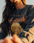 A person dressed in a vintage black "Def Leppard Upcycle Sweatshirt" by People Of Leisure, featuring a graphic leopard design and the word "Pyromania," accentuates their outfit with long hair and hoop earrings. The upcycled fashion is photographed from a low angle, offering a distinctive perspective to the image.