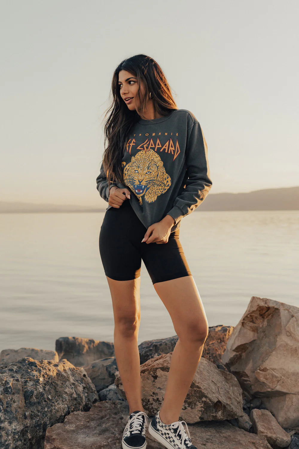 A woman stands on rocks by the water at sunset, wearing a vintage black Def Leppard Upcycle Sweatshirt from People Of Leisure, paired with black shorts and black and white sneakers. She has long hair and gazes off to the side, with a tranquil lake and evening sky serving as her backdrop.