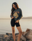 A woman stands on rocks by the water at sunset, wearing a vintage black Def Leppard Upcycle Sweatshirt from People Of Leisure, paired with black shorts and black and white sneakers. She has long hair and gazes off to the side, with a tranquil lake and evening sky serving as her backdrop.