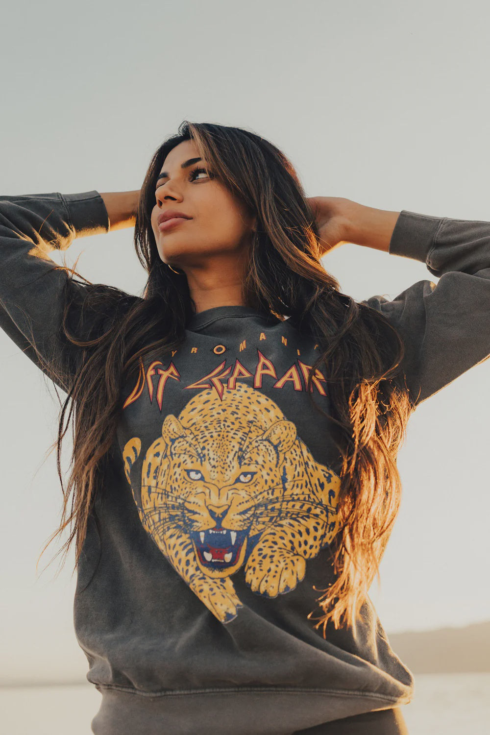 A woman with long hair stands outside, gazing to the side. She's dressed in a vintage Def Leppard Upcycle Sweatshirt by People Of Leisure, which showcases a vibrant tiger graphic and text. The clear sky in the background indicates a sunny day as she raises her hands behind her head.