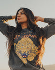 A woman with long hair stands outside, gazing to the side. She's dressed in a vintage Def Leppard Upcycle Sweatshirt by People Of Leisure, which showcases a vibrant tiger graphic and text. The clear sky in the background indicates a sunny day as she raises her hands behind her head.