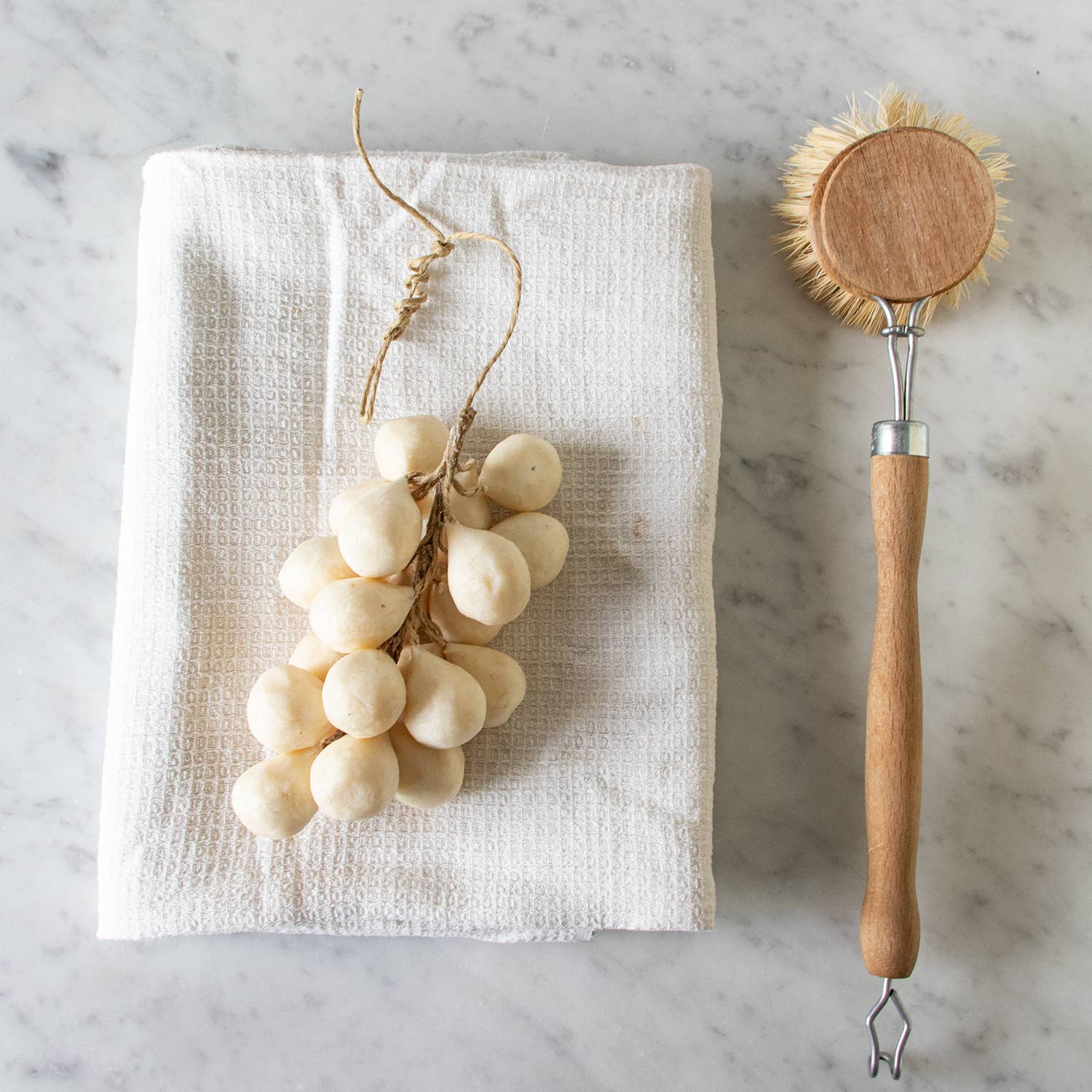 A bar of Faire Handcrafted Grapes Soap sits on a folded white cloth beside a wooden dish brush with bristles, all arranged on a marble surface.