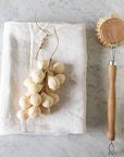 A bar of Faire Handcrafted Grapes Soap sits on a folded white cloth beside a wooden dish brush with bristles, all arranged on a marble surface.
