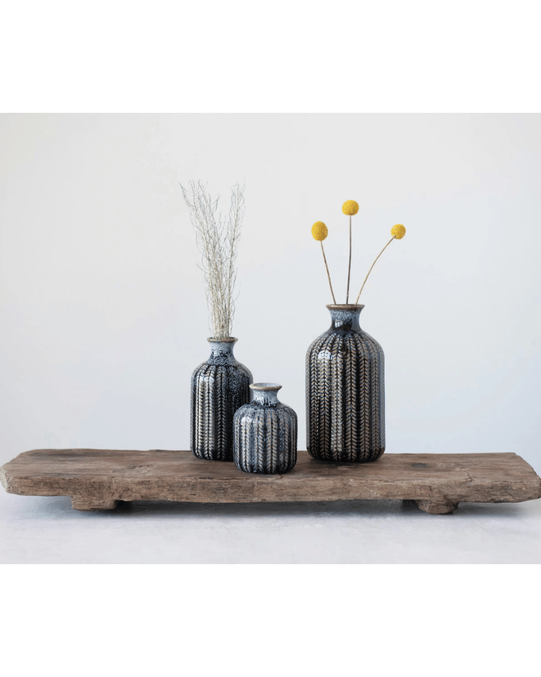 Three Creative Co-op stoneware vases, finished with a reactive glaze and adorned with blue and black patterns, sit on a rustic wooden plank. One holds tall reeds, another cradles yellow billy ball flowers, all set against a minimalist white wall backdrop.