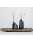 Three Creative Co-op stoneware vases, finished with a reactive glaze and adorned with blue and black patterns, sit on a rustic wooden plank. One holds tall reeds, another cradles yellow billy ball flowers, all set against a minimalist white wall backdrop.