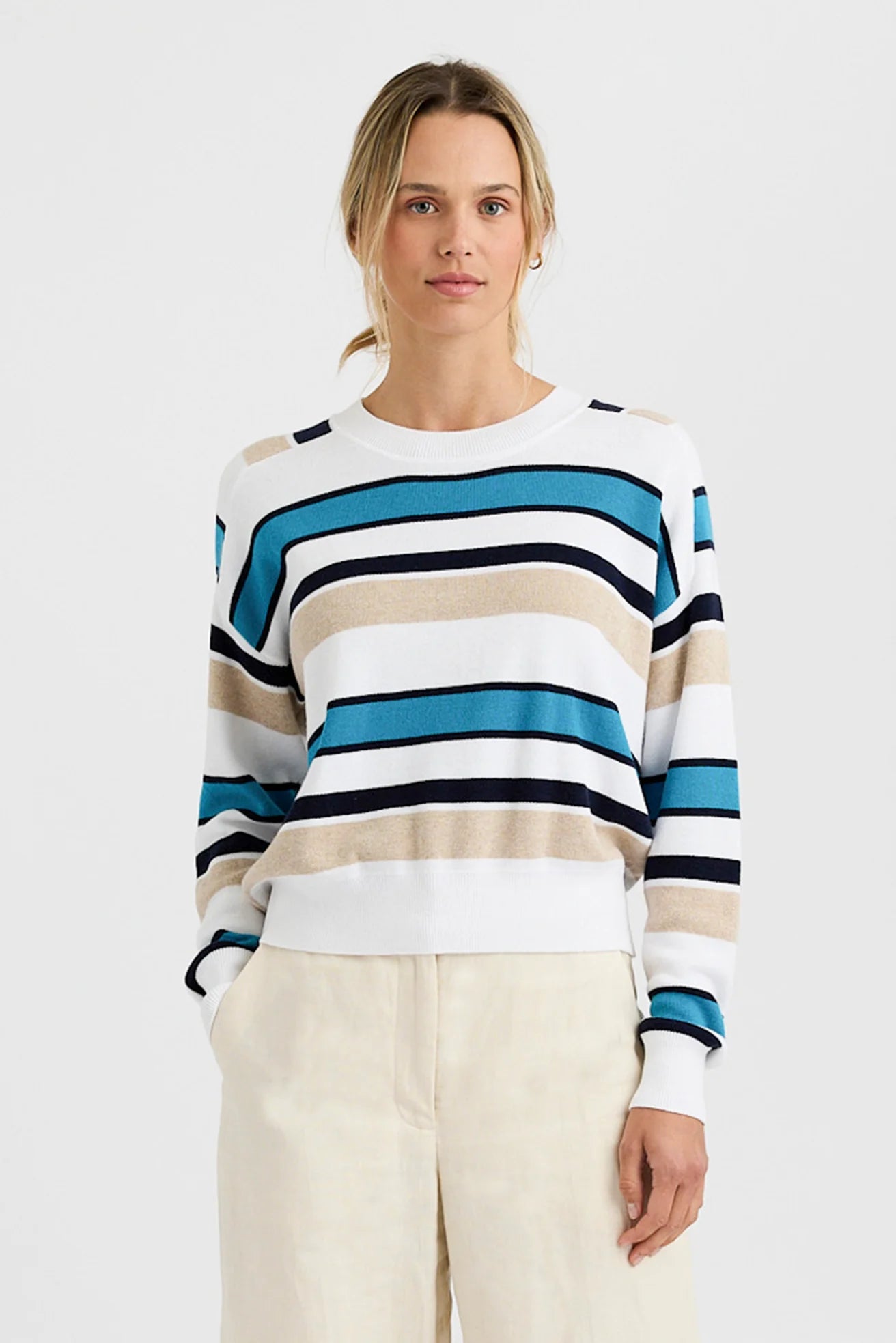 A person stands against a plain background, wearing the One Grey Day Warwick Pullover featuring blue, beige, white, and navy stripes, paired with light-colored Warwick shorts and hands in their pockets.
