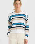 A person stands against a plain background, wearing the One Grey Day Warwick Pullover featuring blue, beige, white, and navy stripes, paired with light-colored Warwick shorts and hands in their pockets.