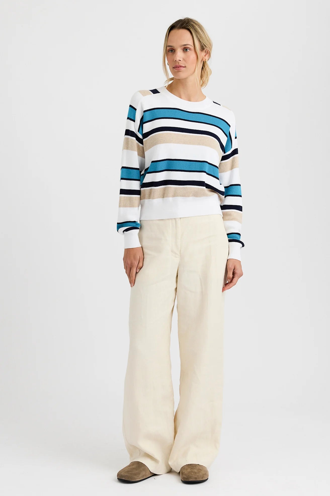 A person stands against a plain background, wearing the One Grey Day Warwick Pullover—a blue, white, and tan striped cotton-blend top with dorito stitching—paired with wide-leg cream pants and brown shoes.