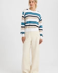 A person stands against a plain background, wearing the One Grey Day Warwick Pullover—a blue, white, and tan striped cotton-blend top with dorito stitching—paired with wide-leg cream pants and brown shoes.