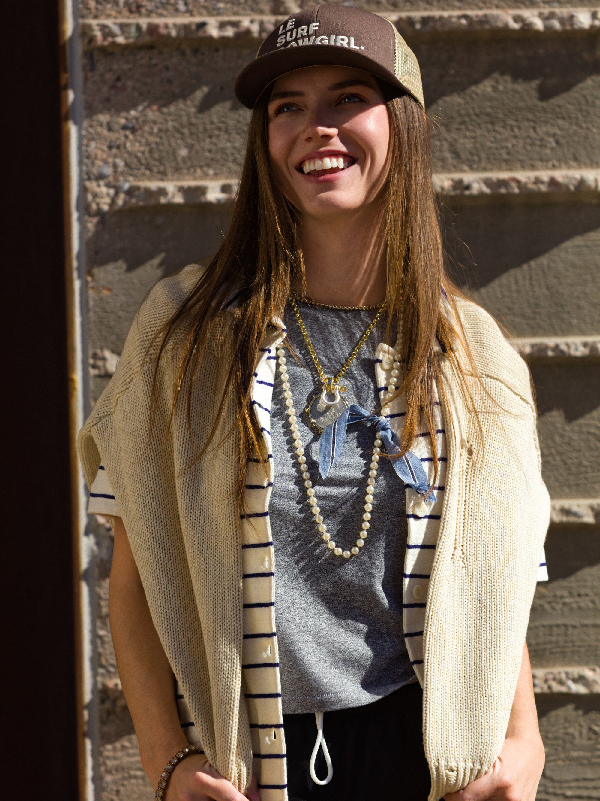 A young woman wearing a brown Ping Pong Surf Club Le Surf Cowgirl Trucker smiles in sunlight, paired with a striped shirt, draped sweater, layered necklaces, and a gray tee while standing against a textured stone wall.
