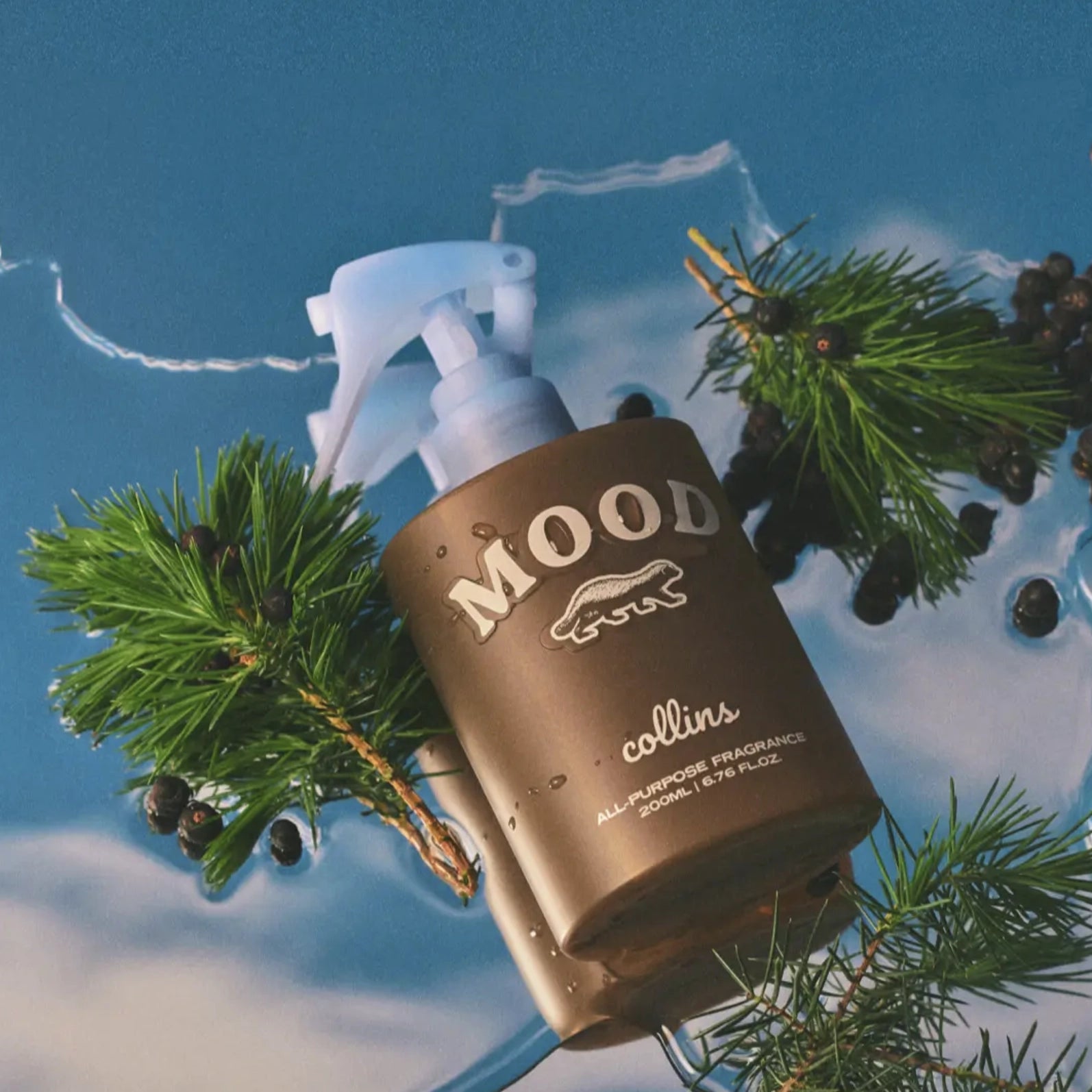 A brown spray bottle labeled "Faire Scottish Juniper Room Spray" sits among green pine branches and dark berries on a blue, watery surface, emphasizing its ethanol-free formula.