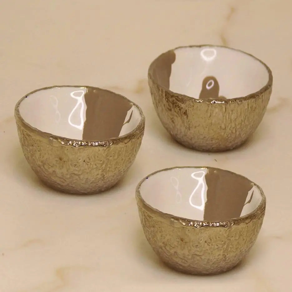 Three small Dansbury Snack Bowls by Faire, artisan crafted with textured metallic gold exteriors and smooth glossy white interiors, are arranged on a light beige surface.