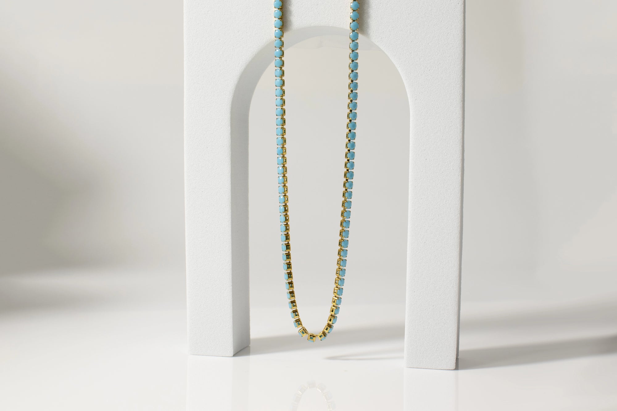 The Faire Turquoise Tennis Necklace, featuring 18K Gold PVD coated stainless steel and turquoise rhinestones, is displayed draped over a white arch-shaped stand on a glossy surface with a soft neutral background.