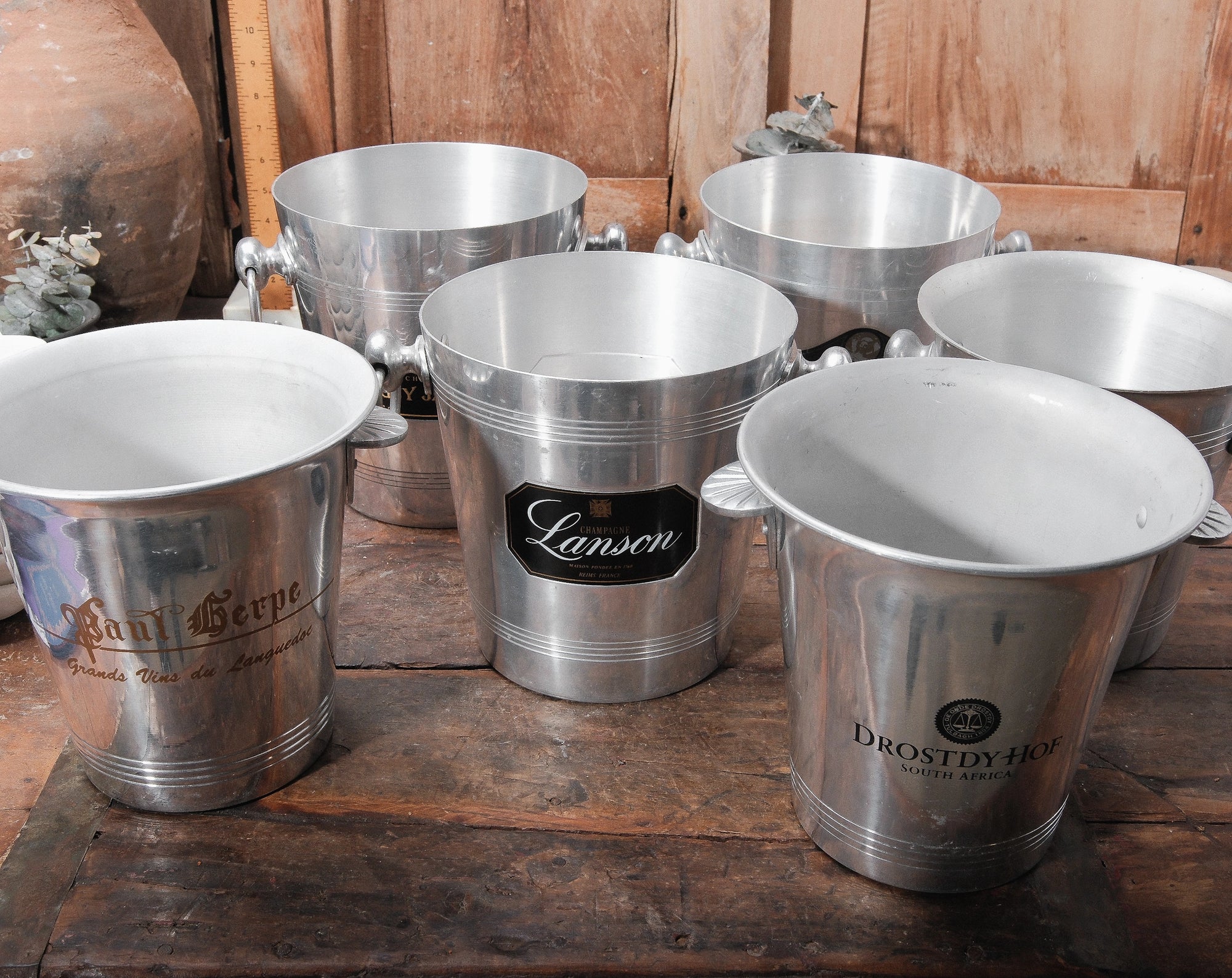 Five Go Home Vintage Champagne Buckets, upcycled and featuring unique logos and inscriptions, are arranged on a wooden surface with warm wooden paneling in the background.