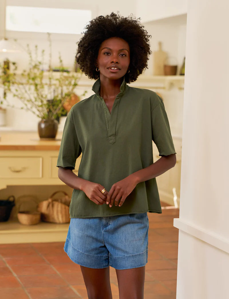 A woman with curly hair wears the Frank & Eileen Patty Popover SS Henley in olive green and denim shorts, standing in a bright kitchen with plants and baskets in the background.
