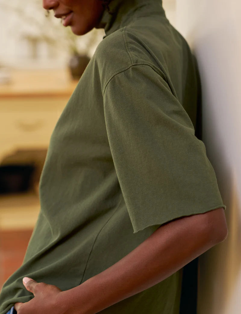 Someone in a loose-fitting, short sleeve Patty Popover SS Henley by Frank & Eileen in olive green leans against a light wall, with the focus on their upper body and arm and a softly blurred indoor background.