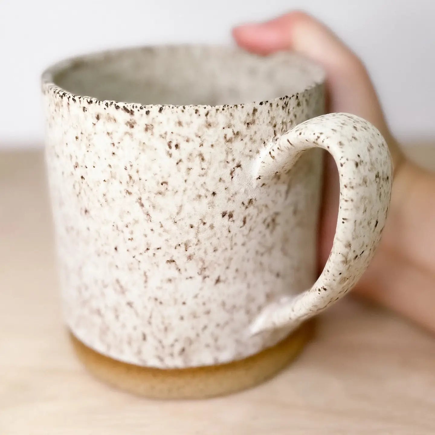 A hand holds Faire’s Large Speckle Mug, an 18oz hand-thrown ceramic with a curved handle, a textured off-white surface with brown speckles, and a brown base, placed on a light wooden surface.