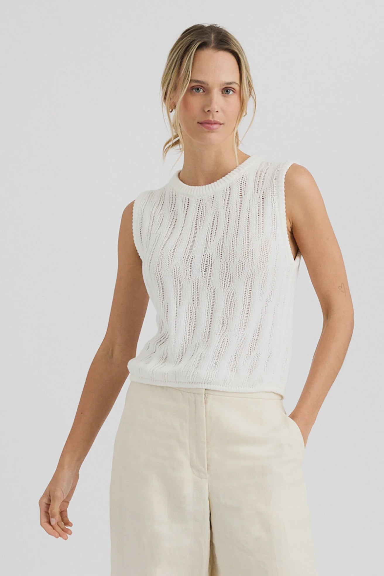 A woman in the One Grey Day Carrick Tank, an airy knit sleeveless white cotton top, paired with light wide-leg pants, stands against a plain light background, looking forward with a relaxed expression.