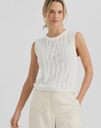 A woman in the One Grey Day Carrick Tank, an airy knit sleeveless white cotton top, paired with light wide-leg pants, stands against a plain light background, looking forward with a relaxed expression.