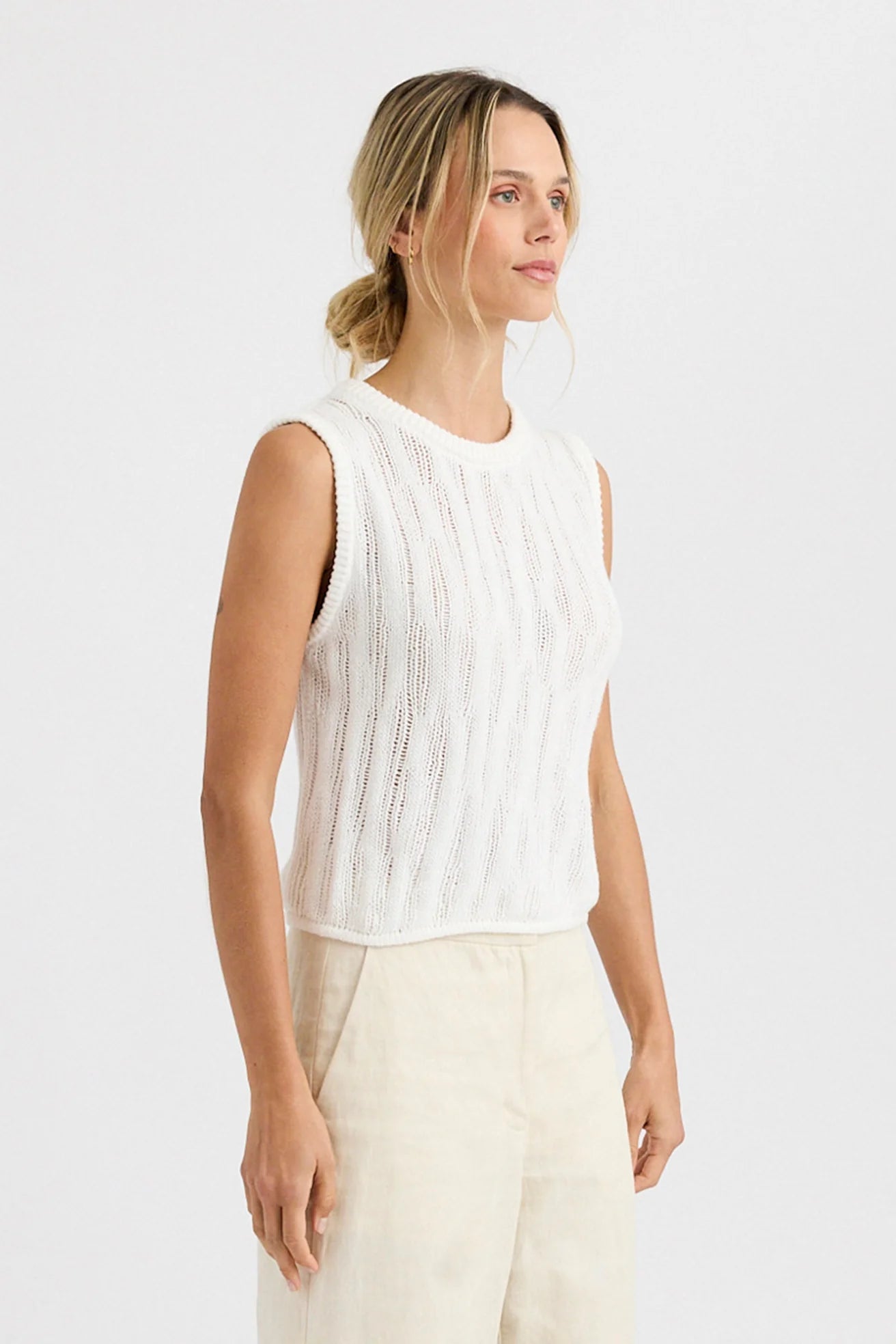 A woman with blonde hair in a loose bun wears the Carrick Tank by One Grey Day, a relaxed fit, sleeveless white knit top, paired with light beige pants. She stands against a plain white background and gazes slightly to the side.