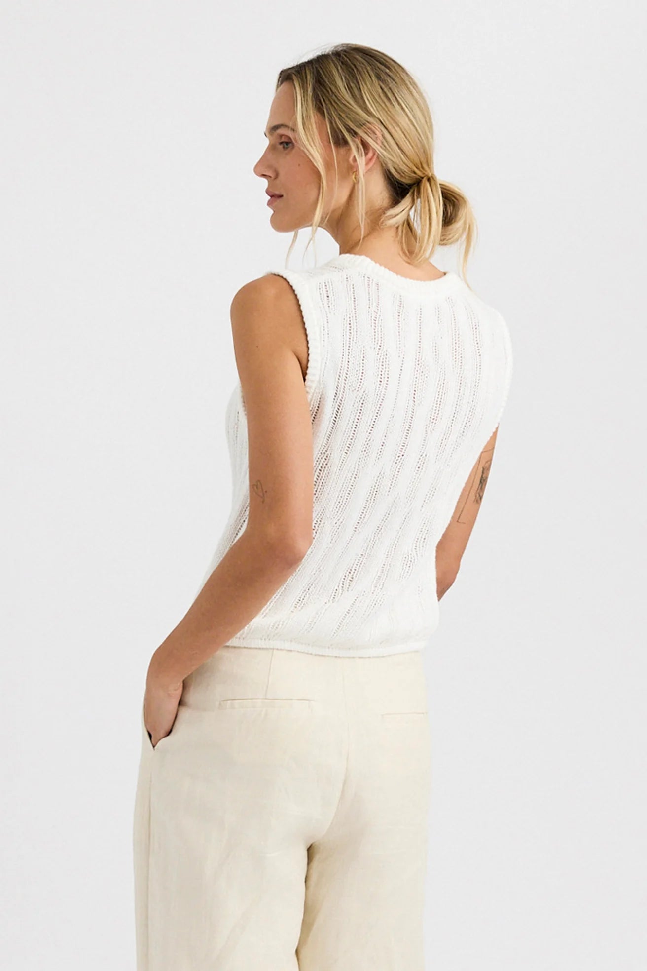 A woman with blonde hair in a low ponytail wears the One Grey Day Carrick Tank, a relaxed fit sleeveless white knit top, paired with light beige pants, standing with her back slightly turned and hands in her pockets against a white background.