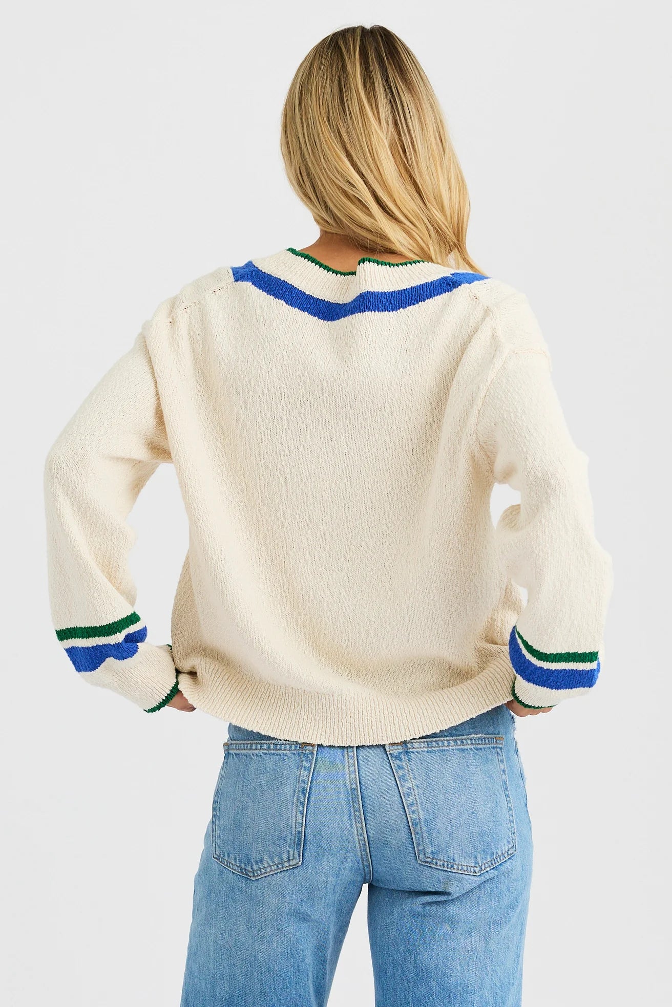 A woman with long blonde hair stands with her back to the camera, wearing the One Grey Day Venus V Neck sweater featuring blue and green striped details, paired with light blue jeans.