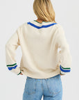 A woman with long blonde hair stands with her back to the camera, wearing the One Grey Day Venus V Neck sweater featuring blue and green striped details, paired with light blue jeans.