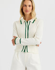 A woman with long blonde hair stands with arms crossed, wearing the One Grey Day Royale Button Up cardigan in white with green trim and white high-waisted shorts against a plain light background.