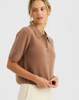 A woman stands against a plain background, looking left with a neutral expression, wearing the One Grey Day Summer Cashmere Polo in light brown and cream-colored pants.