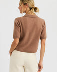 A woman with blonde hair in a low bun wears the One Grey Day Summer Cashmere Polo in light brown and cream pants, standing with her back to the camera against a plain white background.