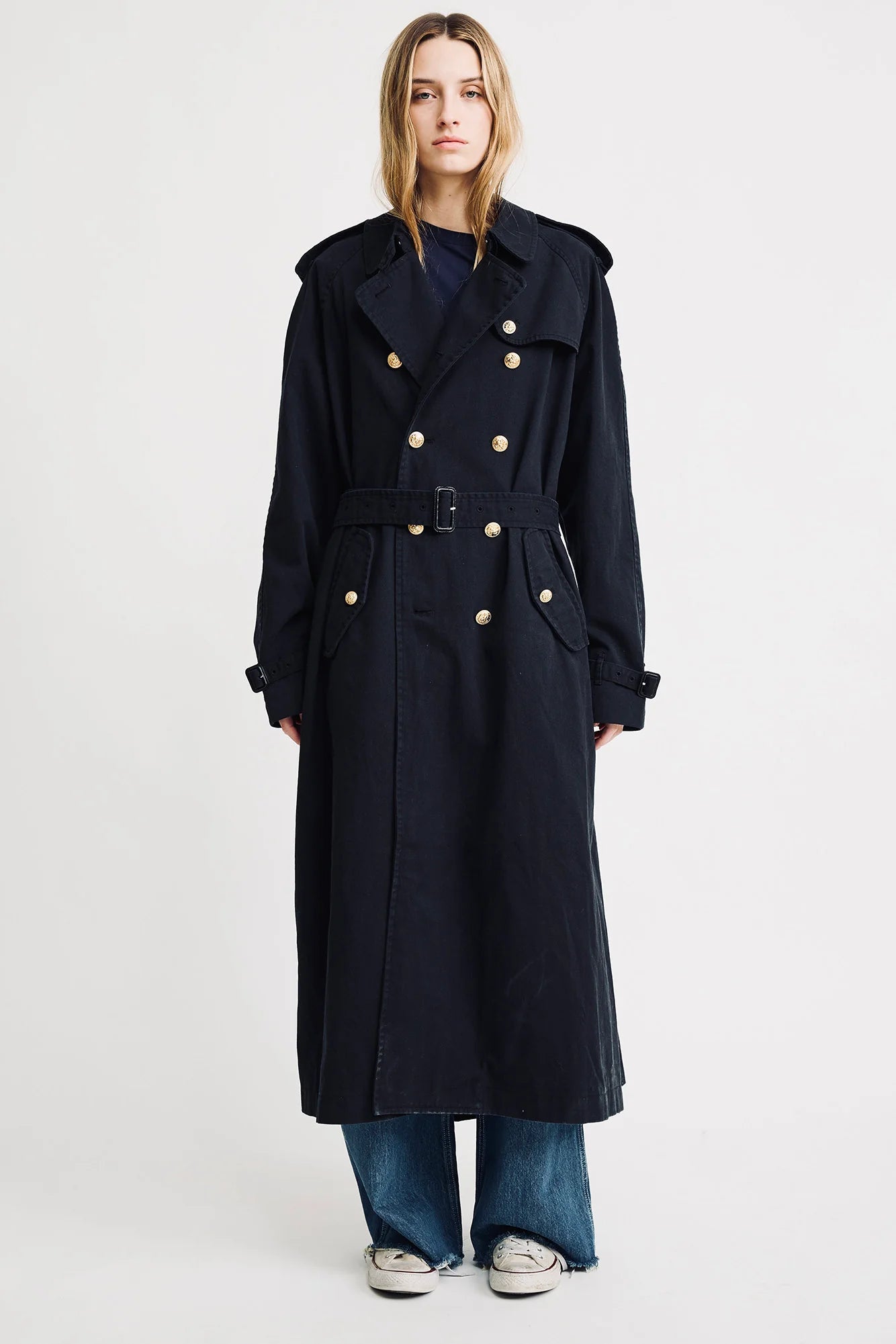 A person stands facing forward against a plain white background, wearing Denimist's OVERSIZED TRENCH COAT in black with gold-tone buttons, paired with blue jeans and white sneakers.