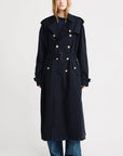 A person stands facing forward against a plain white background, wearing Denimist's OVERSIZED TRENCH COAT in black with gold-tone buttons, paired with blue jeans and white sneakers.
