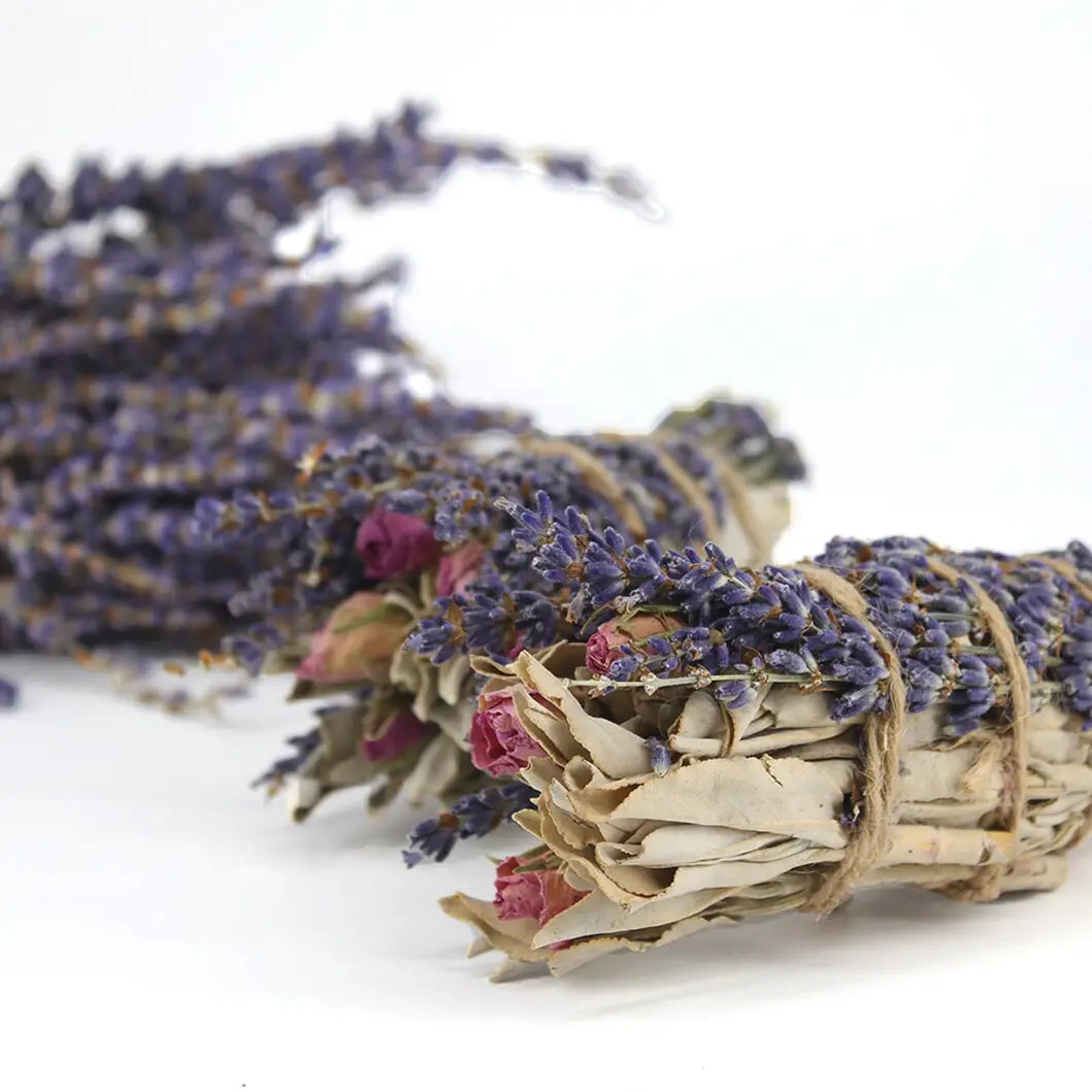 The Faire Lavender, Sage and Rose Smudge features dried sage, lavender, and small red rosebuds tied with twine—ideal for spiritual cleansing as a smudge stick—displayed on a white surface with another bundle softly blurred behind.