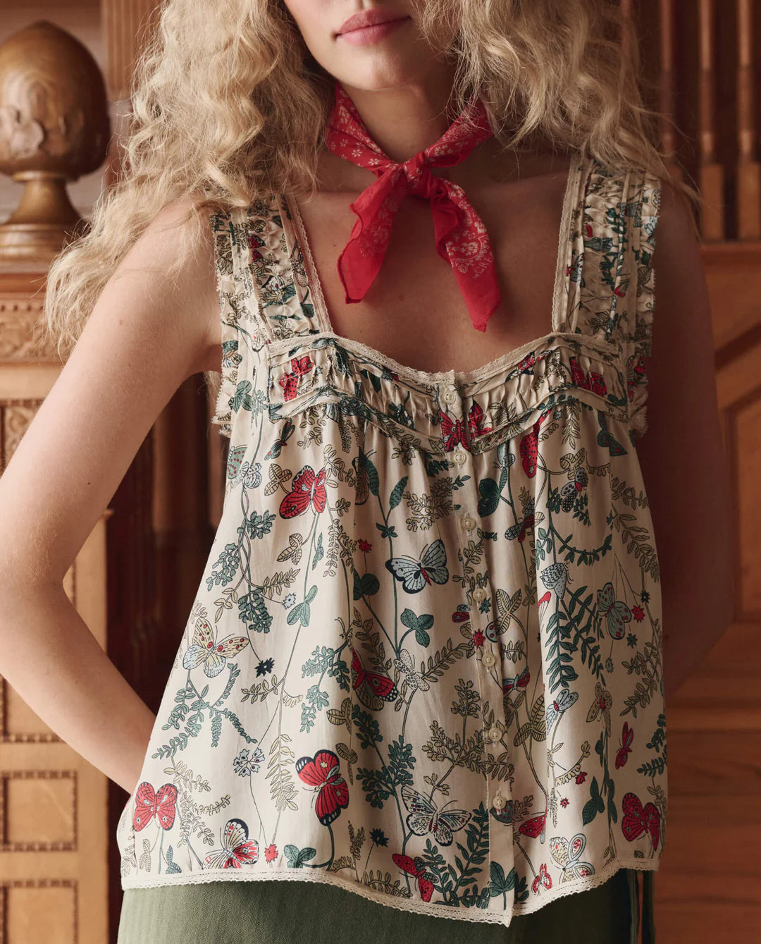 A woman with wavy blonde hair wears The Victorian Pleat Tank by The Great Inc. in vintage rose print, paired with a red bandana around her neck. She stands indoors near ornate wooden furniture.