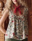 A woman with wavy blonde hair wears The Victorian Pleat Tank by The Great Inc. in vintage rose print, paired with a red bandana around her neck. She stands indoors near ornate wooden furniture.